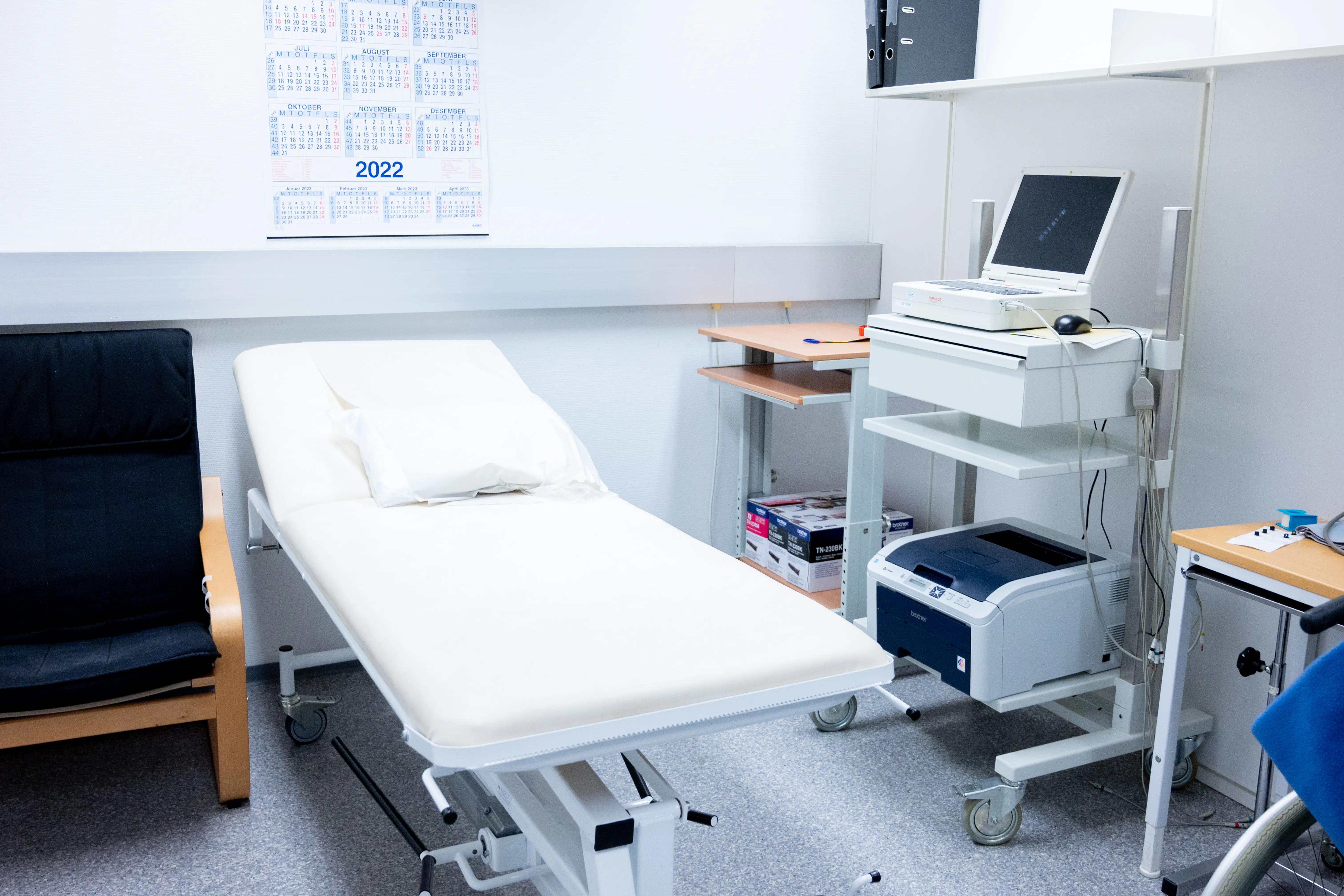 Foto av et undersøkelsesrom med stol, seng, apparat og benker.